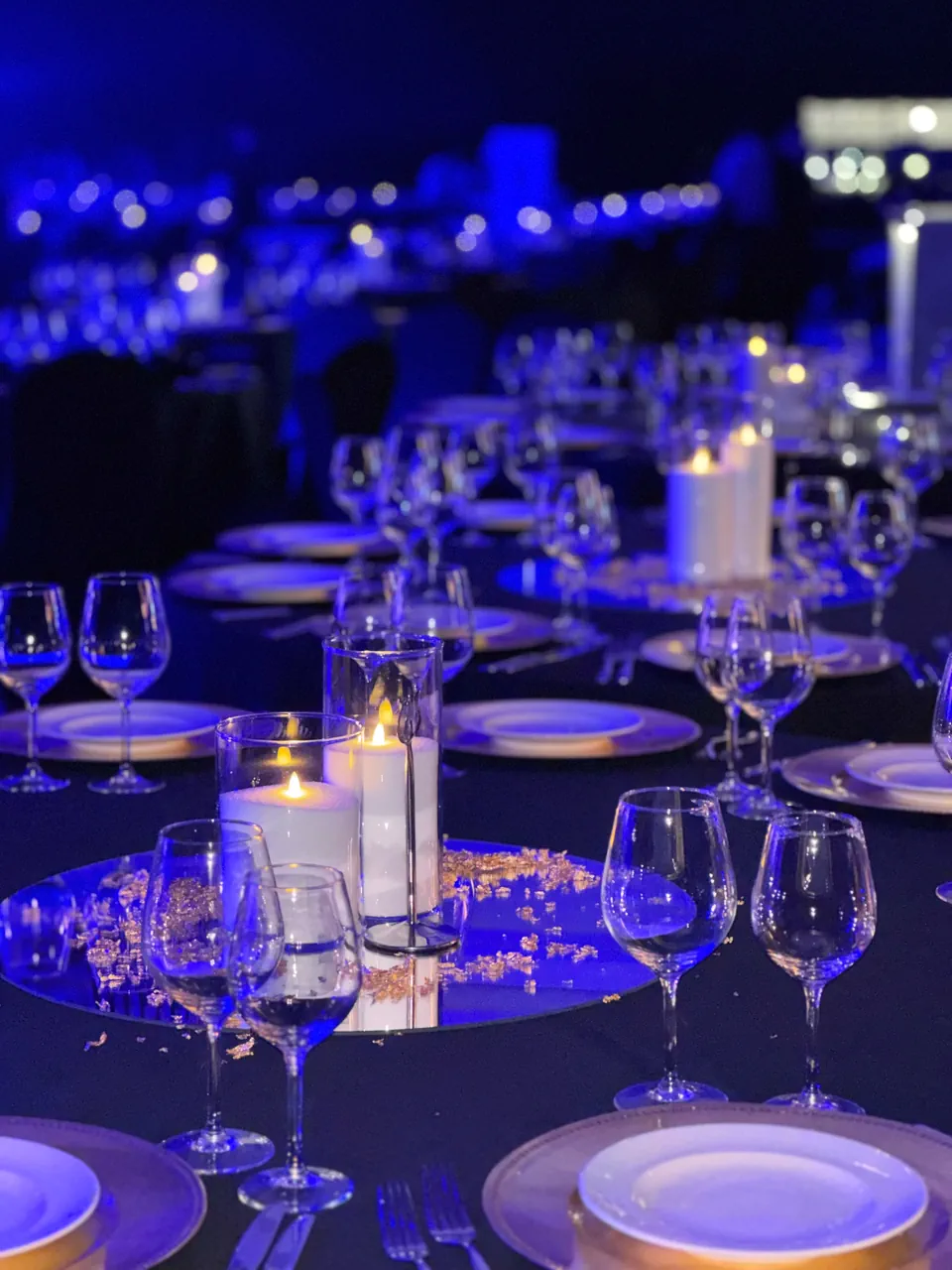 Para w szarych marynarkach i jeansach pozuje w sali z drewnianą podłogą i ceglanym sufitem, z dekoracjami z wiszących białych kwiatów, zasłon i czarnej lampy