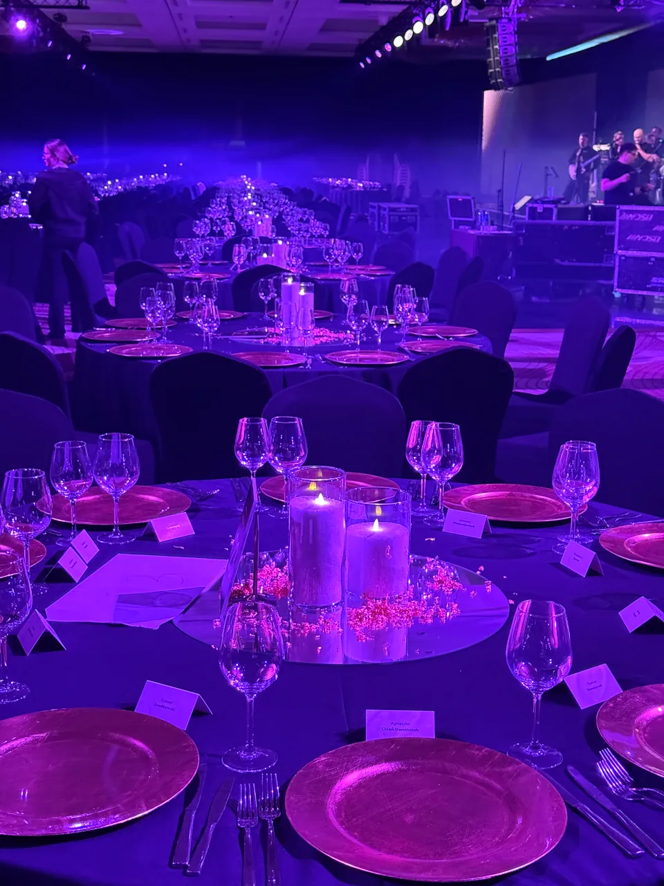 Okrągłe stoły bankietowe nakryte czarnymi obrusami, ozdobione złotymi podtalerzami i winietkami z nazwiskami gości. Na środku każdego stołu znajduje się lustrzana podstawka z dwiema dużymi świecami w szklanych cylindrach, otoczona dekoracyjnymi kryształkami. Cała sala jest spowita intensywnym, fioletowym światłem, a w tle widać scenę przygotowaną do występu