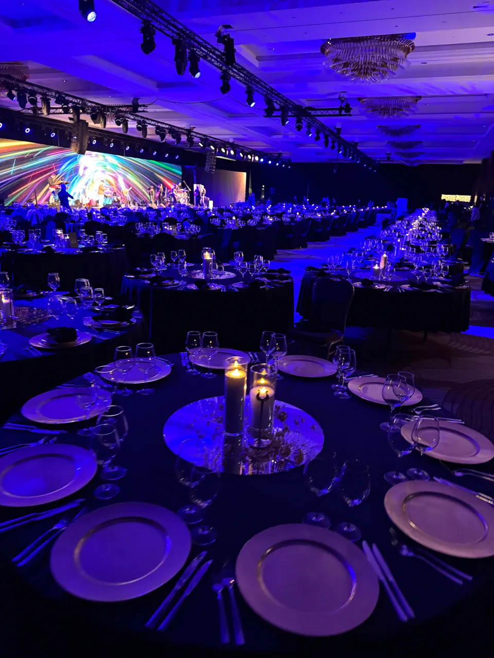 Widok z góry na elegancko zastawiony okrągły stół bankietowy podświetlony na fioletowo. Na środku znajduje się lustrzana taca ze świecami, a dookoła rozmieszczono srebrzyste talerze i kieliszki. W oddali widać rzędy podobnych stołów oraz duży ekran multimedialny na scenie, wyświetlający barwną grafikę podczas trwającego wydarzenia