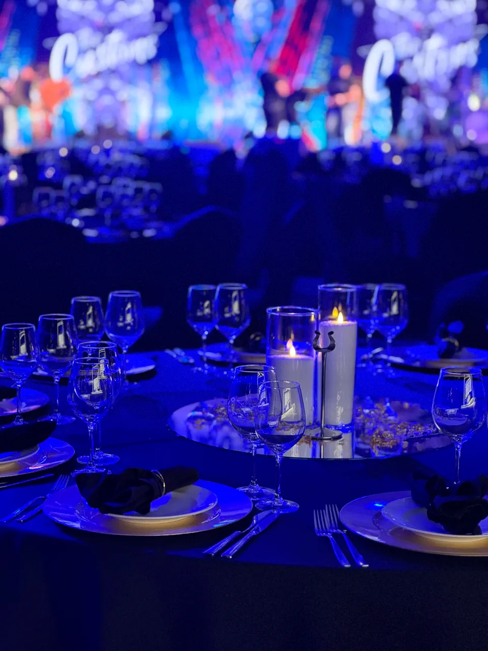 Artystyczne zbliżenie na zastawę stołu bankietowego w intensywnym, niebieskim oświetleniu. Na pierwszym planie widoczne kieliszki do wina oraz talerze z elegancko ułożonymi czarnymi serwetkami. W centrum stołu płoną dwie świece na lustrzanej podstawie, a mocno rozmyte tło ukazuje scenę z dynamicznymi efektami świetlnymi