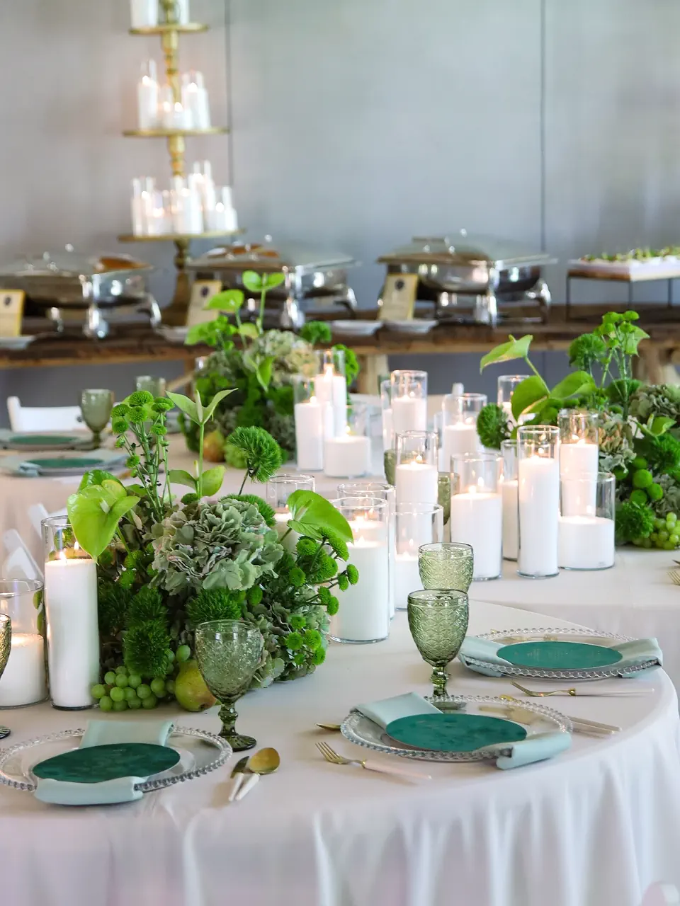 Elegancka dekoracja okrągłego stołu bankietowego w tonacji zieleni i bieli. Na stole znajdują się obfite kompozycje z zielonych kwiatów, hortensji i anturium, liczne białe świece w szklanych cylindrach oraz oliwkowe kieliszki z fakturowanego szkła. W tle widoczny jest bufet z podgrzewaczami i piętrową paterą ze świecami