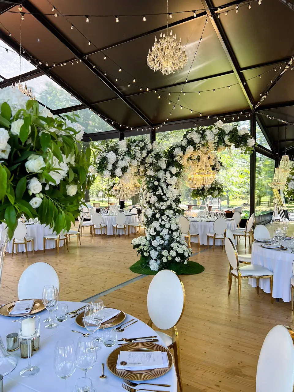 Wnętrze luksusowego namiotu eventowego udekorowanego na wesele, z centralnie umieszczoną monumentalną konstrukcją z białych kwiatów i zwisającymi kryształowymi żyrandolami. W sali ustawiono okrągłe stoły z białymi obrusami, złotą zastawą i eleganckimi krzesłami, a pod sufitem rozpięto sznury dekoracyjnych lampek. Przeszklone ściany namiotu ukazują otaczającą zieleń ogrodu