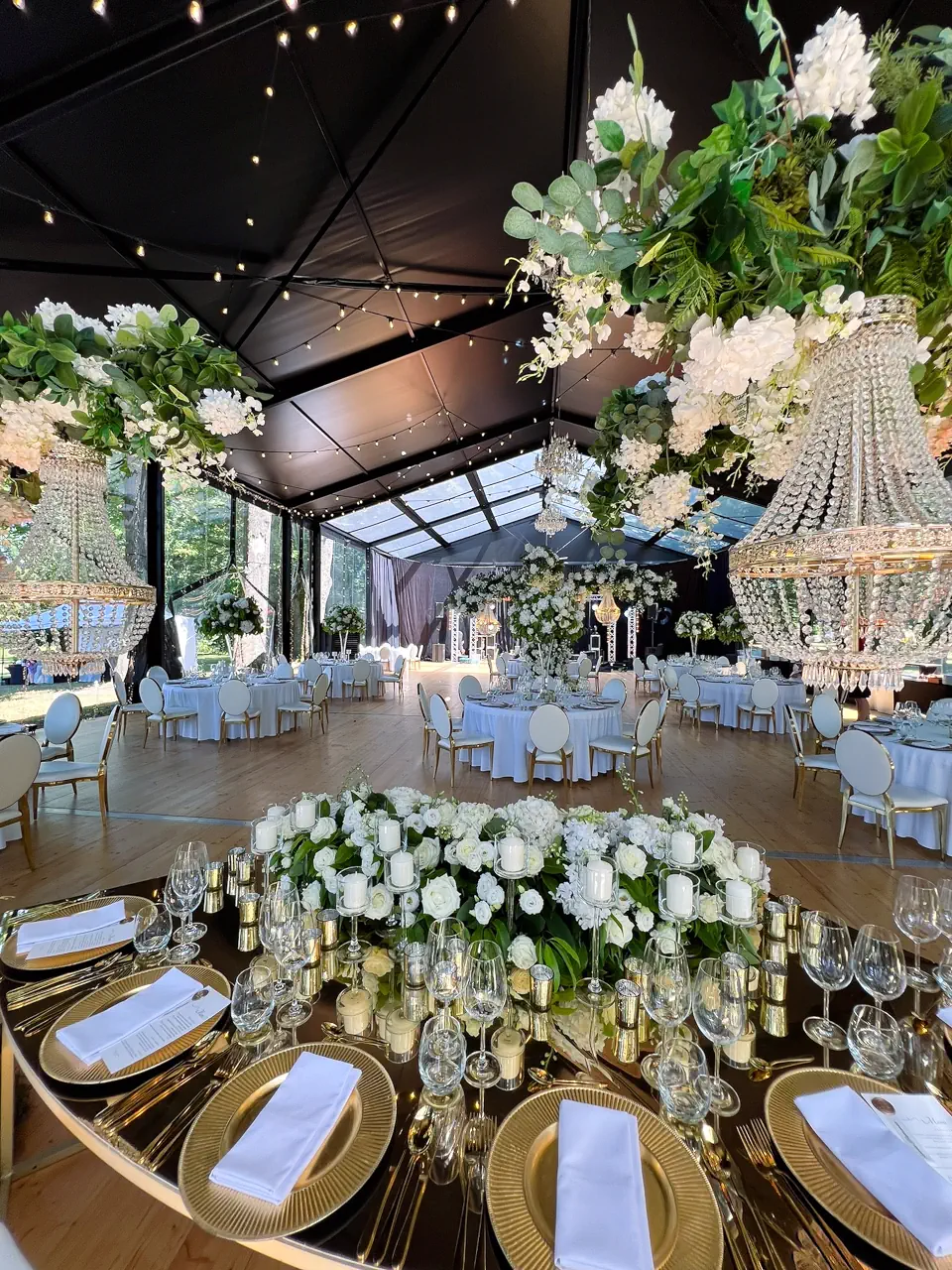 Szerokie ujęcie wnętrza eleganckiego namiotu bankietowego z lustrzanym stołem prezydialnym udekorowanym białymi kwiatami i dziesiątkami świec w szklanych osłonkach. Nad stołami wiszą okazałe kryształowe żyrandole oplecione zielenią, a w głębi sali widać okrągłe stoliki gości pod czarnym poszyciem dachu rozświetlonym setkami drobnych lampek. Całość tworzy luksusową i nastrojową atmosferę wieczornego przyjęcia