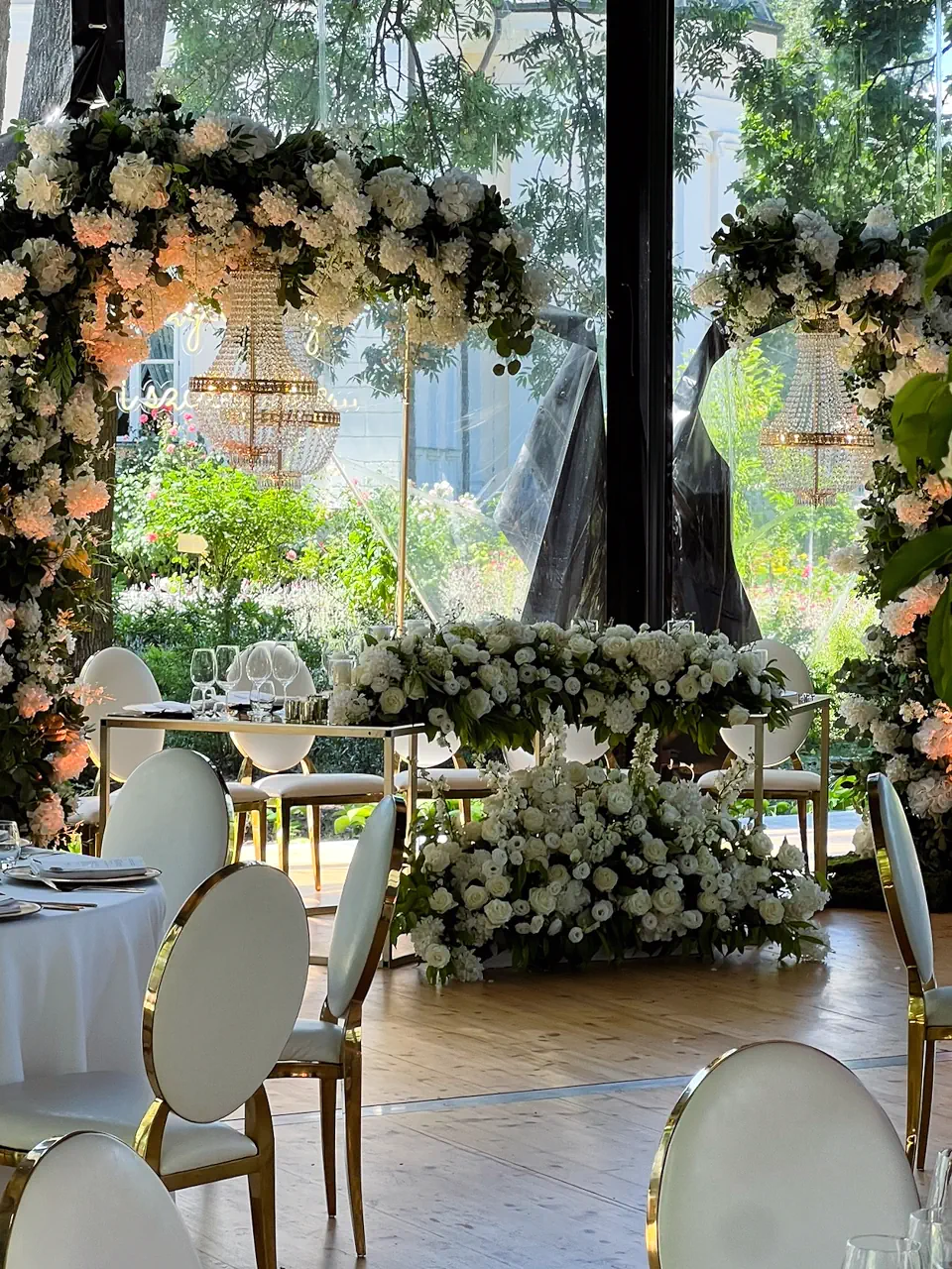 Elegancka dekoracja kwiatowa stołu pary młodej w szklanym namiocie, z obfitą girlandą z białych kwiatów spływającą ze stołu na podłogę. Nad stołem góruje łuk z jasnych kwiatów z podwieszonym kryształowym żyrandolem, a w tle widać przeszkloną ścianę z widokiem na słoneczny ogród. Na pierwszym planie ustawione są nowoczesne biało-złote krzesła bankietowe