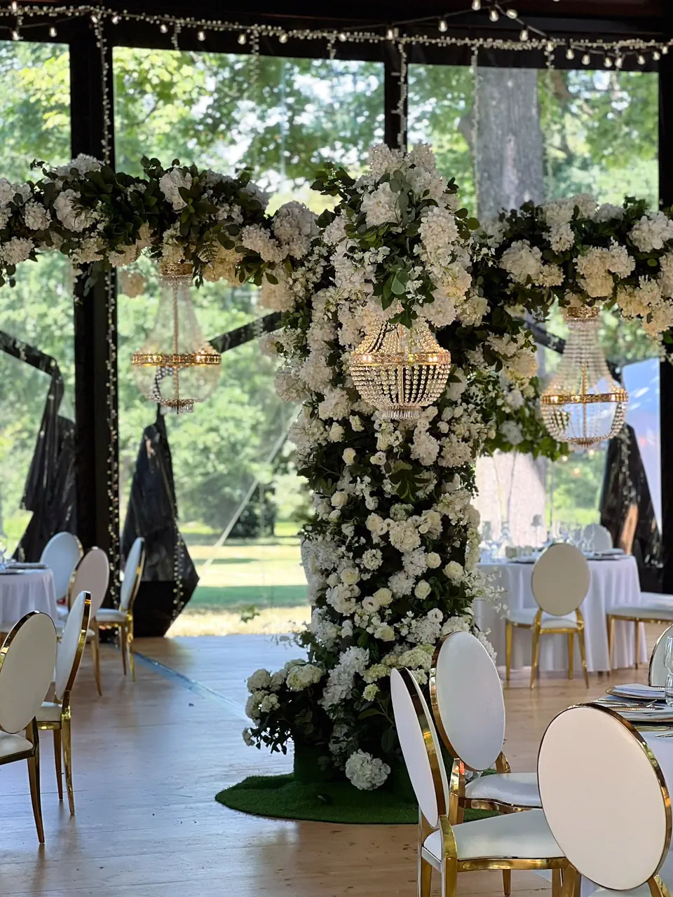 Spektakularna kolumna kwiatowa z białych hortensji i róż, stanowiąca centralny punkt dekoracji w luksusowym namiocie weselnym. Z konstrukcji kwiatowej wyrastają ramiona z podwieszonymi, świecącymi kryształowymi żyrandolami, a w tle widać elegancko nakryte okrągłe stoły i biało-złote krzesła. Przeszklone ściany namiotu wpuszczają naturalne światło i widok na park