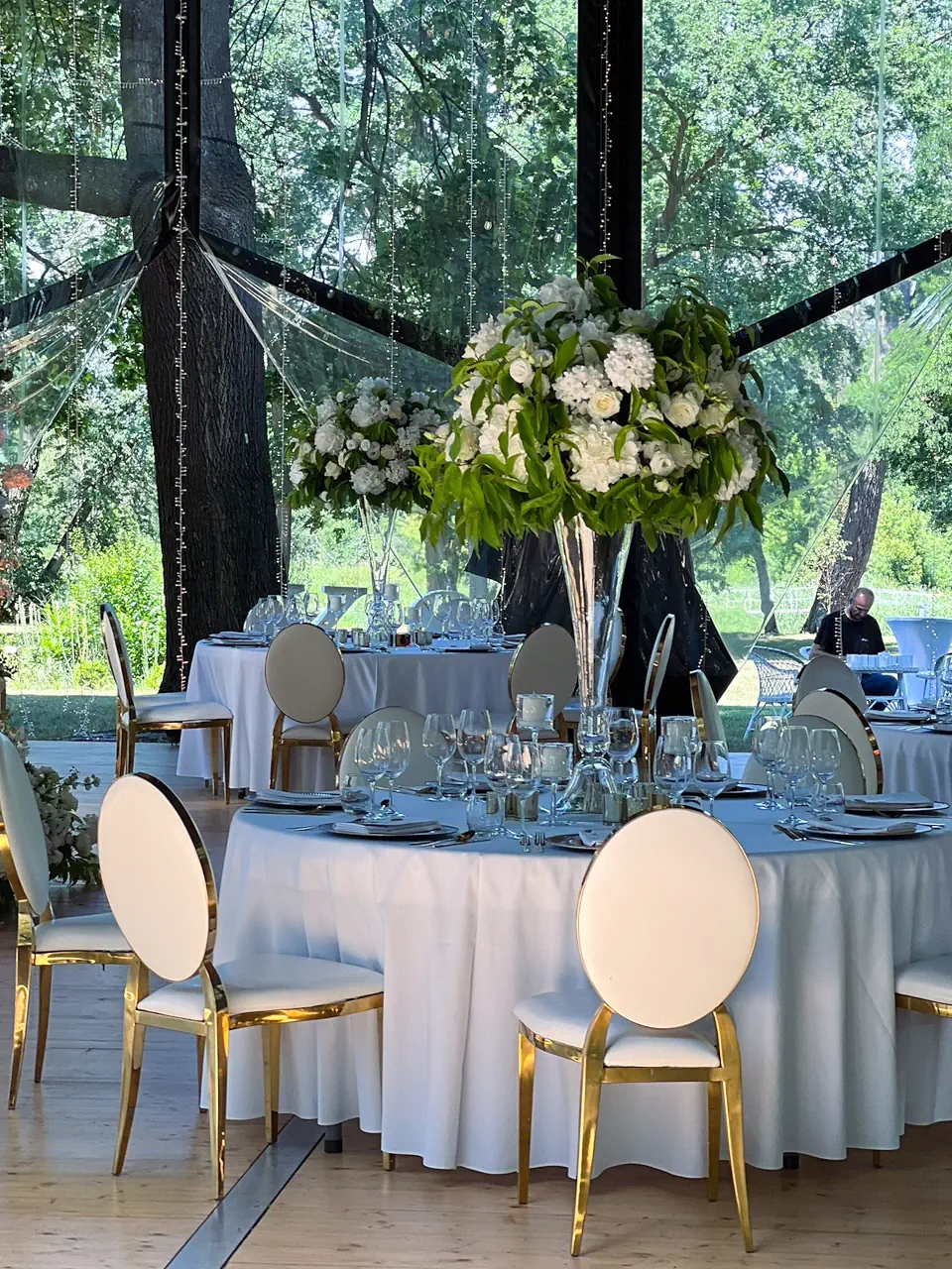 Elegancki wystrój sali weselnej w szklanym namiocie, z okrągłymi stołami przykrytymi białymi obrusami i nowoczesnymi biało-złotymi krzesłami. Na środku stołu stoi wysoki szklany wazon z bogatą kompozycją z białych kwiatów i zielonych liści, a w tle widać pnie drzew i parkową zieleń prześwitującą przez transparentne ściany konstrukcji. Nad całością wiszą delikatne girlandy świetlne
