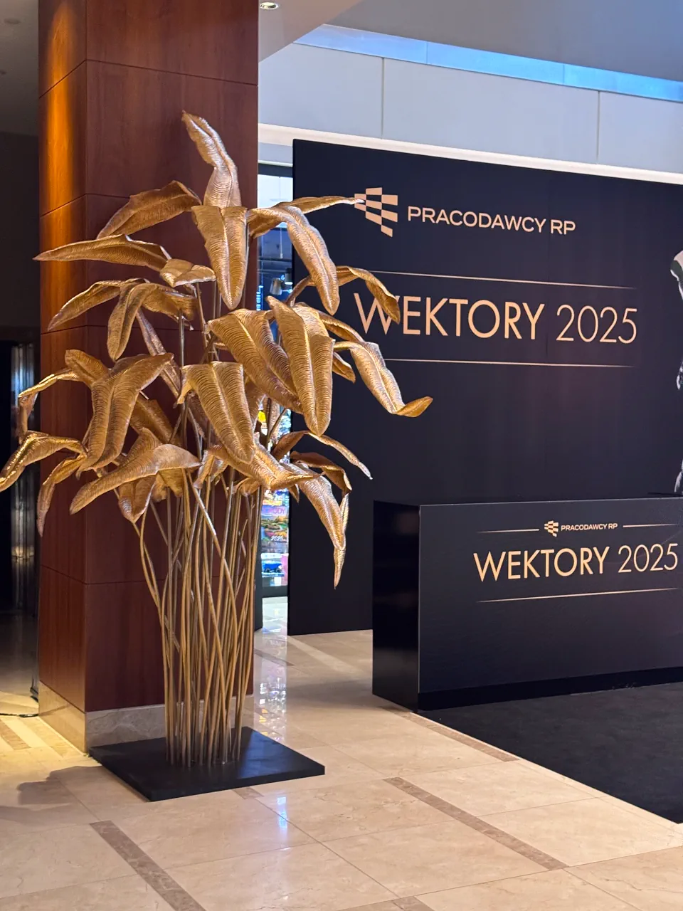 Złota dekoracja florystyczna w formie dużych liści palmowych na wysokich łodygach, ustawiona przy drewnianym filarze. W tle widoczna elegancka czarna ścianka eventowa oraz lada recepcyjna z logotypem Pracodawcy RP i napisem „Wektory 2025”. Nowoczesna aranżacja przestrzeni bankietowej na jasnej, błyszczącej podłodze