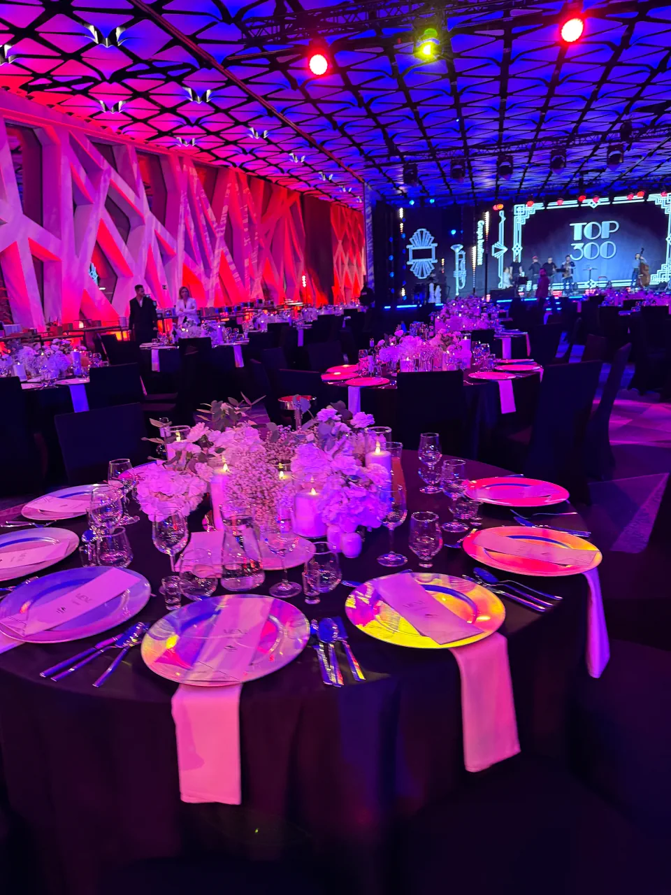 Uroczysta kolacja bankietowa z okazji gali „TOP 300”. Na pierwszym planie okrągły stół z czarnym obrusem, elegancką zastawą i dekoracją z białych kwiatów oraz świec. Cała sala wypełniona jest nastrojowym fioletowo-różowym światłem, a w tle widać scenę z wielkim ekranem i wystrój w stylu Wielkiego Gatsby’ego