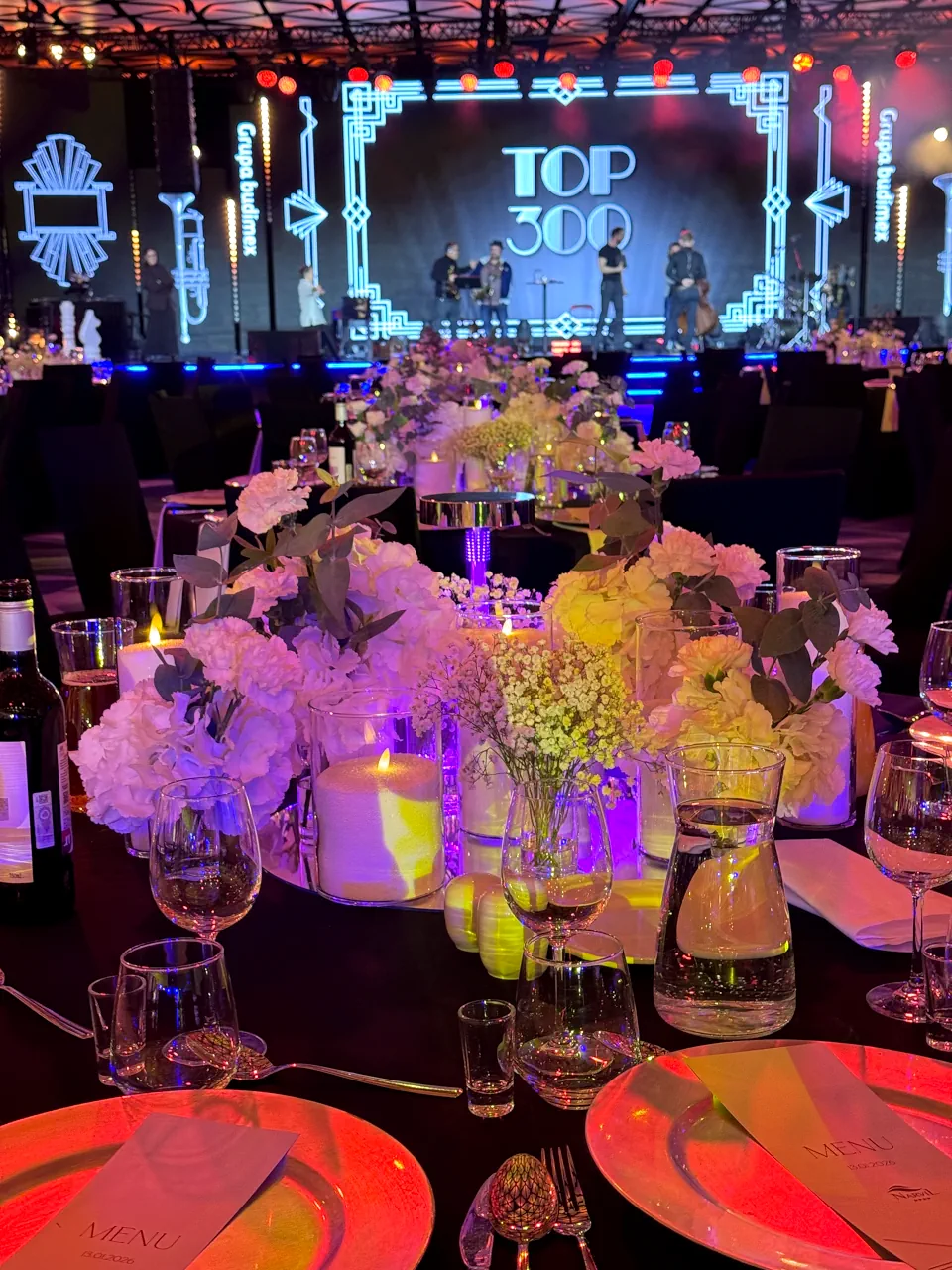 Zbliżenie na dekorację stołu bankietowego podczas gali „TOP 300”. Kompozycja składa się z białych hortensji i goździków, uzupełnionych świecami w szklanych naczyniach, które odbijają fioletowe światło reflektorów. W tle widać scenę z zespołem muzycznym i ekranem w geometrycznym stylu art déco z logotypem „Grupa Budimex”