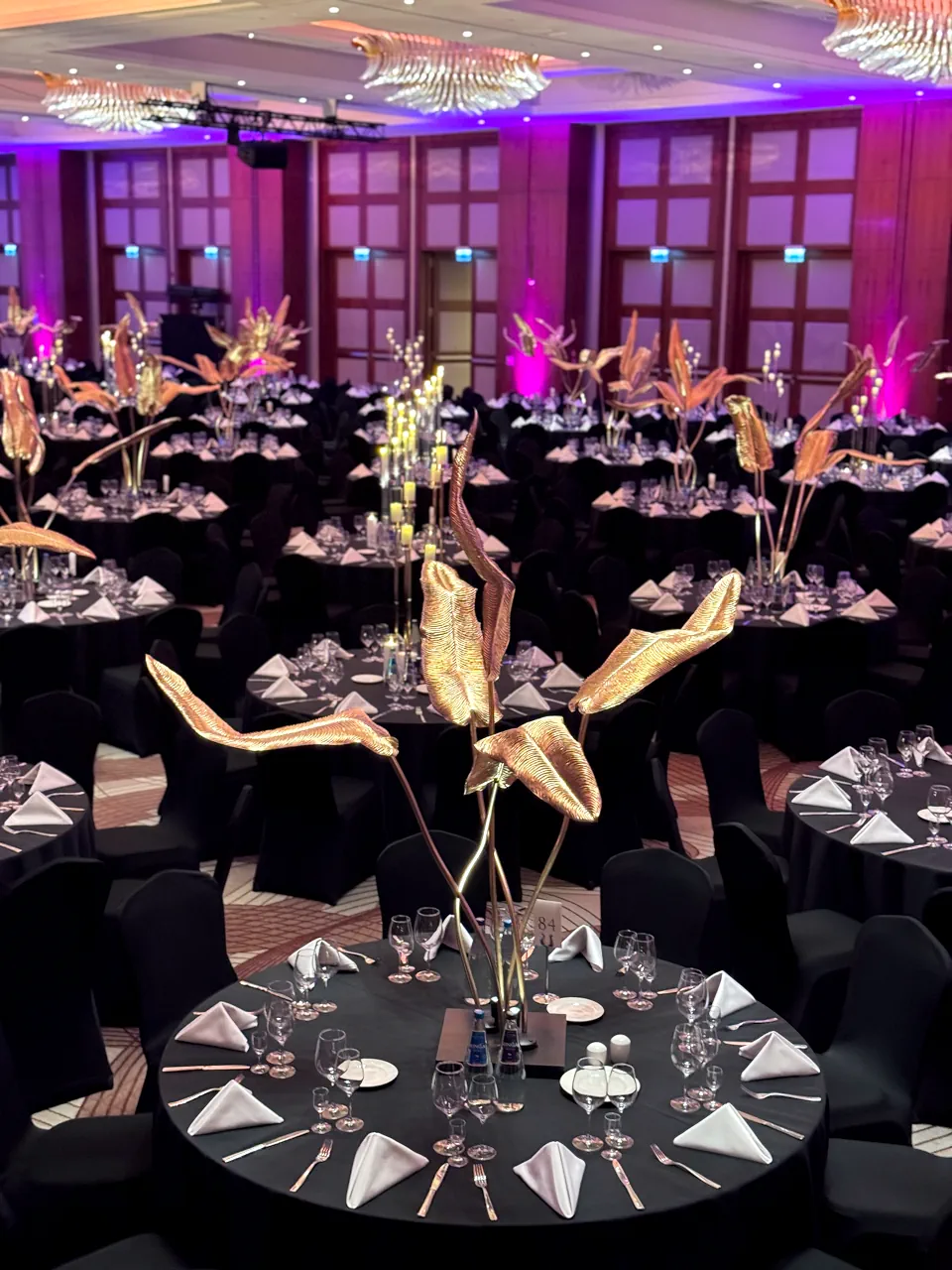 Przestronna sala bankietowa wypełniona licznymi okrągłymi stołami z czarnymi obrusami i białymi serwetkami. Centralnym punktem każdego stołu jest wysoka, złota dekoracja w kształcie abstrakcyjnych liści palmowych. Wnętrze oświetlono intensywnym fioletowym światłem, a sufit zdobią kryształowe, nowoczesne żyrandole, tworząc luksusową atmosferę