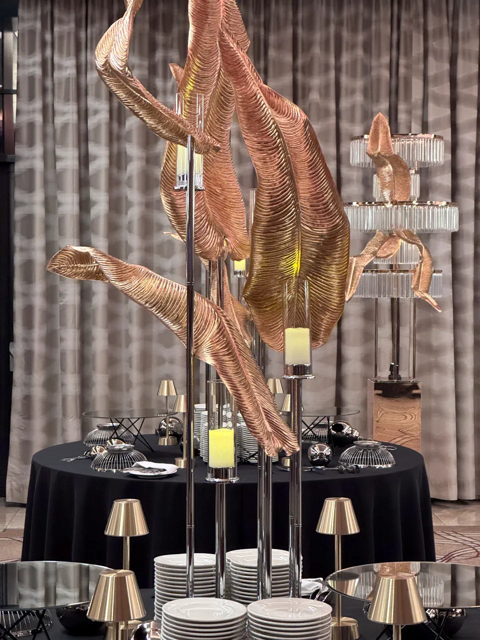 Zbliżenie na artystyczną dekorację bufetu z rzeźbionymi, miedziano-złotymi liśćmi i świecami w szklanych osłonach na wysokich, srebrnych stojakach. Na czarnym obrusie ustawiono stosy białych talerzy oraz nowoczesne złote lampki stołowe. W tle widoczna elegancka kurtyna i kryształowa kolumna świetlna