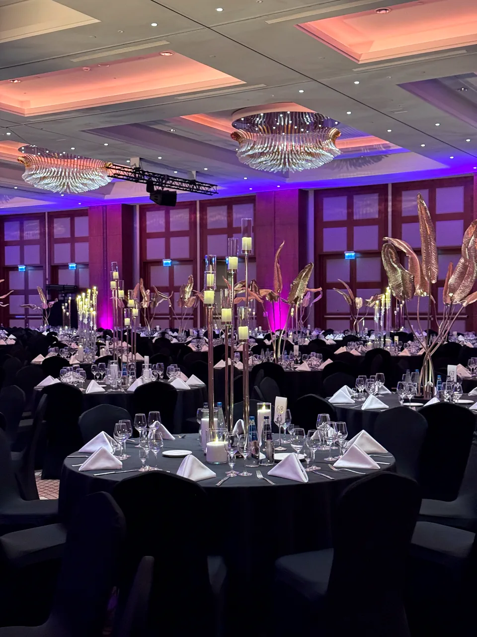 Szerokie ujęcie eleganckiej sali bankietowej z okrągłymi stołami, czarnymi obrusami i białymi serwetkami złożonymi w trójkąty. Na stołach znajdują się wysokie świeczniki z płonącymi świecami oraz złote dekoracje florystyczne. Sufit zdobią efektowne, rozłożyste żyrandole, a ściany i wnęki są podświetlone nastrojowym fioletowym i pomarańczowym światłem