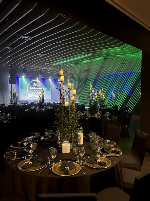 Elegancka dekoracja eventowa na gali w Warszawie, okrągły stół z świecami i zielenią, stoły z ciemnymi obrusami i złotymi akcentami, scena z nowoczesnym oświetleniem