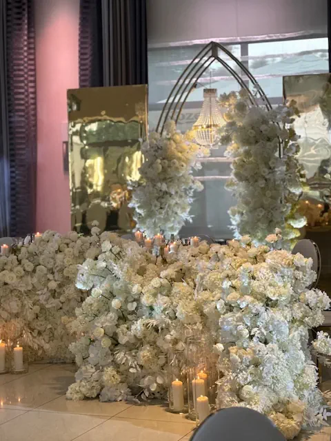 Luksusowa dekoracja sali weselnej w hotelu Rubin Radom w eleganckim, białym stylu. Główne tło stanowi ściana z białych hortensji, róż i orchidei, ozdobiona licznymi świecami w szklanych naczyniach, które dodają romantycznego blasku. Za dekoracją ustawiono złote lustra i metalową konstrukcję w kształcie łuku, udekorowaną wiszącymi białymi kwiatami. W odbiciu widać imponujący kryształowy żyrandol, podkreślający luksusowy charakter wnętrza. Całość tworzy bajkową, elegancką atmosferę, idealną na ekskluzywne wesele