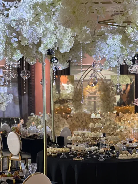 Luksusowa aranżacja sali weselnej w hotelu Rubin Radom w eleganckim, biało-złotym stylu. W centrum znajduje się spektakularna dekoracja sufitowa z białych hortensji i kryształowych zawieszek, które odbijają światło. Poniżej ustawiono bogato zastawiony stół z eleganckimi deserami, w tym piętrowymi paterami z babeczkami i słodkimi przekąskami. W tle złote lustra i biała, kwiatowa ścianka dodają przestrzeni ekskluzywnego charakteru. Białe, tapicerowane krzesła i złote detale dopełniają luksusową atmosferę, idealną na eleganckie wesele lub wyjątkową uroczystość