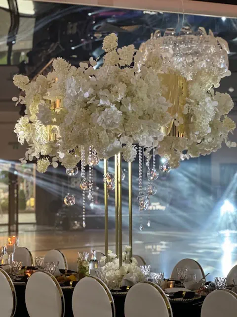 Ekskluzywna dekoracja stołu bankietowego w hotelu Rubin Radom w eleganckim, biało-złotym stylu. Centralnym elementem jest imponująca kompozycja kwiatowa z białych hortensji i orchidei, umieszczona na złotej konstrukcji. Z kompozycji zwisają kryształowe girlandy i szklane kule, odbijające światło, co dodaje luksusowego blasku. W tle reflektory tworzą efektowne świetlne smugi, podkreślając wyjątkową atmosferę wydarzenia. Białe, tapicerowane krzesła z złotymi ramami oraz elegancka zastawa stołowa dopełniają wyrafinowany charakter aranżacji, idealnej na wesele lub ekskluzywną galę
