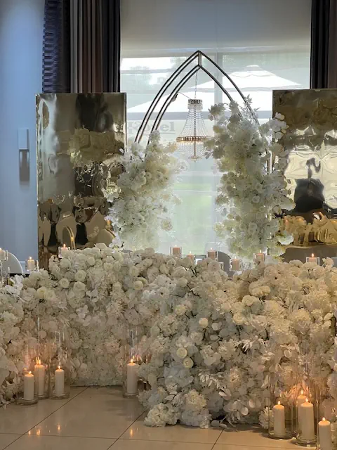 Elegancka dekoracja weselna w luksusowym, biało-złotym stylu. Dominującym elementem jest ściana w hotelu Rubin Radom z gęstej kompozycji białych kwiatów, takich jak róże, hortensje i orchidee, przeplatana wysokimi, szklanymi cylindrami ze świecami, które dodają romantycznego blasku. W tle złote, lustrzane panele odbijają światło, a centralnym punktem jest złoty, metalowy łuk ozdobiony zwisającymi białymi kwiatami. Za nim widoczny jest kryształowy żyrandol, podkreślający ekskluzywny charakter aranżacji. Całość tworzy bajkową scenerię, idealną na luksusowe wesele