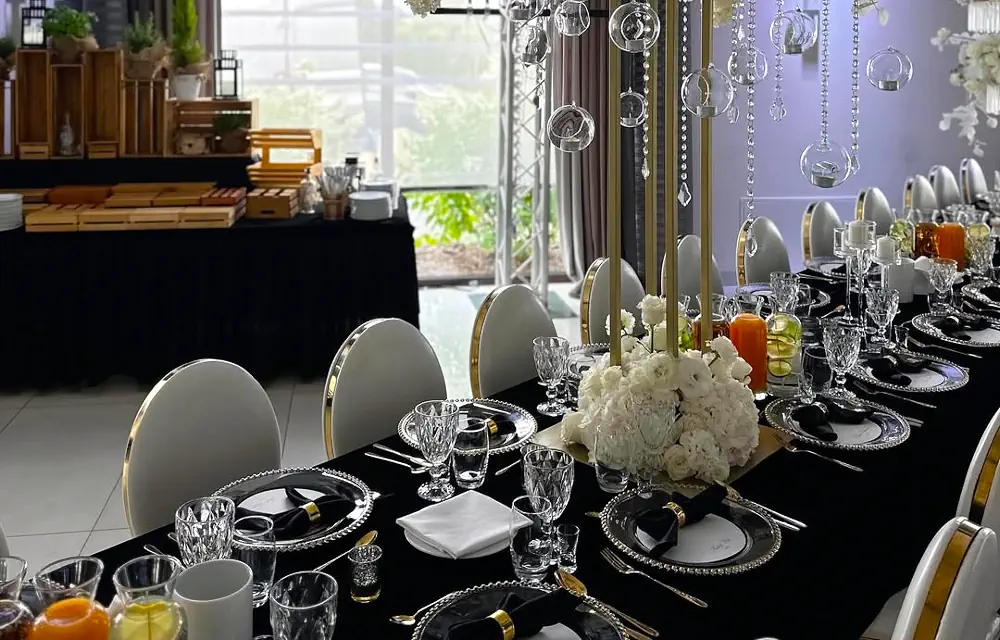 Elegancka aranżacja stołu bankietowego w hotelu Rubin Radom w luksusowym, czarno-złotym stylu. Stół nakryty czarnym obrusem ozdobiono kryształową zastawą, srebrnymi sztućcami i dekoracyjnymi talerzami z perłowym zdobieniem. Na każdym nakryciu ułożono czarne serwetki z ozdobnymi złotymi pierścieniami. Centralnym elementem dekoracji są białe kwiatowe kompozycje u podstawy złotych stojaków, z których zwisają szklane kule i kryształowe girlandy. Pośród świeczników znajdują się bursztynowe świece dodające ciepłego blasku. W tle widać bufet w rustykalnym stylu, tworzący kontrast z eleganckim wystrojem stołu