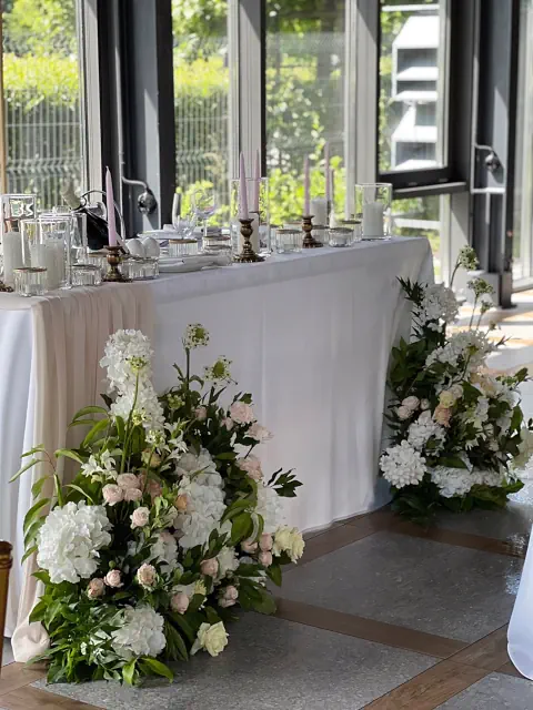 Elegancka dekoracja stołu prezydialnego w Pałacu Mała Wieś w stylu romantycznym. Stół pokryty białym obrusem ozdobiony jest delikatnie opadającą tkaniną w odcieniu jasnego beżu. Na blacie ustawiono kryształowe szkło, smukłe świeczniki z różowymi świecami oraz złote akcenty, dodające wyrafinowanego charakteru. Przy podstawie stołu znajdują się bujne kompozycje kwiatowe z białych hortensji, róż, eustom i zielonych liści, które harmonijnie komponują się z otoczeniem. W tle duże okna wpuszczają naturalne światło, podkreślając lekkość i elegancję wystroju