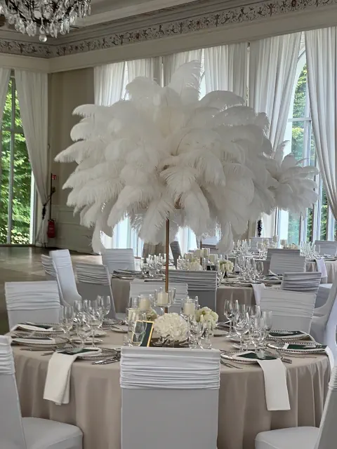 Luksusowa aranżacja sali weselnej w Pałacu Mała Wieś w stylu glamour z eleganckimi dekoracjami. Okrągłe stoły nakryte beżowymi obrusami ozdobione są wysokimi kompozycjami z białych strusich piór na smukłych złotych stojakach, nadając wnętrzu ekstrawaganckiego charakteru w stylu lat 20. XX wieku. Stylowe nakrycia obejmują kryształowe kieliszki, srebrne sztućce i białe serwetki starannie ułożone na talerzach. Krzesła pokryte białymi pokrowcami harmonizują z jasnym, klasycznym wystrojem sali z wysokimi oknami, długimi zasłonami i kryształowym żyrandolem. Całość tworzy wytworną atmosferę idealną na ekskluzywne przyjęcie weselne