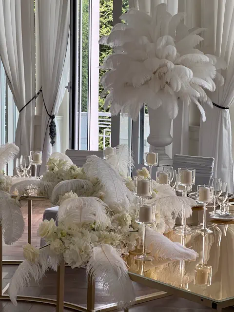Ekskluzywna aranżacja weselna w stylu glamour inspirowanym latami 20. XX wieku. Dekorację stołu stanowią luksusowe kompozycje z białych strusich piór oraz hortensji, rozmieszczone na złotym, lustrzanym blacie. Wysokie, eleganckie świeczniki ze smukłymi białymi świecami dodają romantycznego blasku. W tle białe krzesła z pokrowcami harmonizują z przestronnym wnętrzem Pałacu Mała Wieś o wysokich oknach i zwiewnych zasłonach. Całość tworzy niezwykle elegancką, ponadczasową atmosferę, idealną na luksusowe wesele w stylu art déco