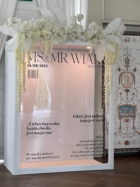 Elegancka dekoracja weselna w formie personalizowanej ścianki przypominającej okładkę magazynu. Konstrukcja w białej ramie zawiera duże, lustrzane tło z nadrukiem „MS & MR WIĄT WEDDING” oraz datą ślubu 26/08/2023. Na powierzchni znajdują się romantyczne cytaty o miłości, dodające uroczystego charakteru. Górna część dekoracji ozdobiona jest białymi hortensjami, zwisającą glicynią oraz strusimi piórami, nadając całości luksusowego stylu glamour. Tło z odbiciem wnętrza dodaje głębi, tworząc efektowne miejsce do zdjęć na weselu