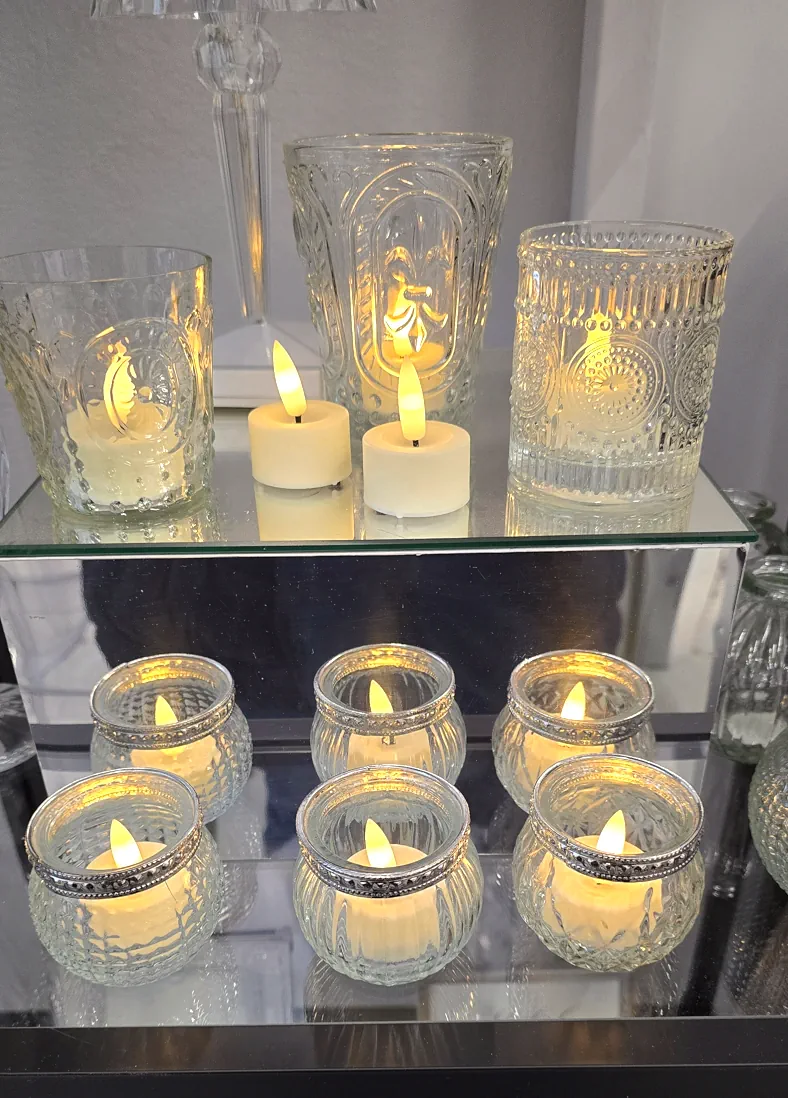 Zestaw świec LED tealight w różnych szklanych kloszach o zdobionych wzorach, ustawionych na lustrzanej półce, tworzących ciepłe i eleganckie oświetlenie