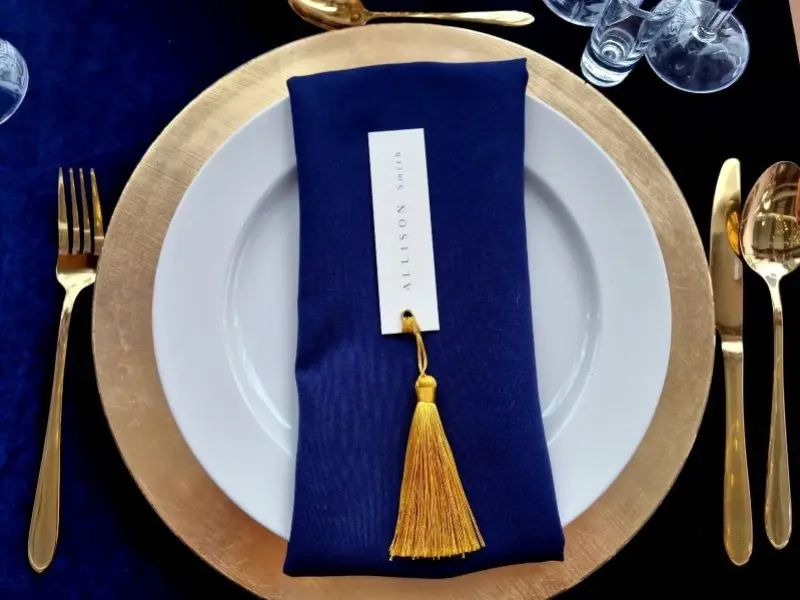 Eleganckie nakrycie stołu z białym talerzem na złotej podkładce. Na talerzu znajduje się granatowa serwetka, złożona i ozdobiona złotym frędzlem, a na niej zawieszona karta z imieniem „ALLISON SMITH”. Po lewej stronie talerza leży złoty widelec, a po prawej złota łyżka i nóż. W tle widoczne są kieliszki oraz ciemnoniebieski obrus, co nadaje całej aranżacji elegancki i wyrafinowany charakter.