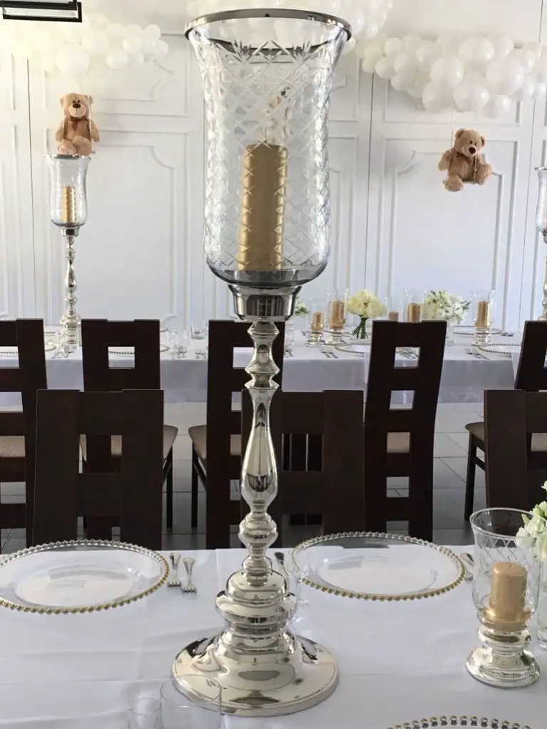 Elegancka dekoracja stołu z wysokim, srebrnym świecznikiem z ozdobnym, szlifowanym szklanym kloszem, wewnątrz którego znajduje się złota świeca. Świecznik ma klasyczną, połyskującą podstawę z dekoracyjnym trzonem, dodając całości luksusowego charakteru. Stół nakryty jest białym obrusem i uzupełniony przezroczystymi talerzami z ozdobnym złotym rantem oraz kieliszkami. W tle widoczne są balonowe dekoracje w odcieniach bieli oraz pluszowe misie zawieszone na ścianie, co sugeruje przyjęcie o charakterze uroczystym, np. chrzest lub baby shower