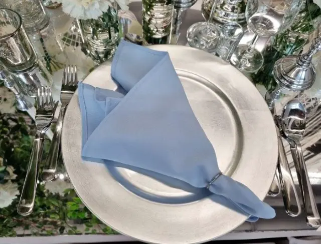Elegancko nakryty stół z talerzem w kolorze srebrnym, na którym leży błękitna serwetka złożona w elegancki trójkąt i spięta srebrnym pierścieniem. Obok talerza widoczne są sztućce: widelec po lewej stronie talerza, nóż i łyżka po prawej stronie. W tle widać przezroczyste kieliszki i wysokie szklanki, które dodają blasku aranżacji. Stół ozdobiony jest subtelnymi, kwiatowymi akcentami, a wszystko razem tworzy elegancką, spójną kompozycję idealną na uroczystości