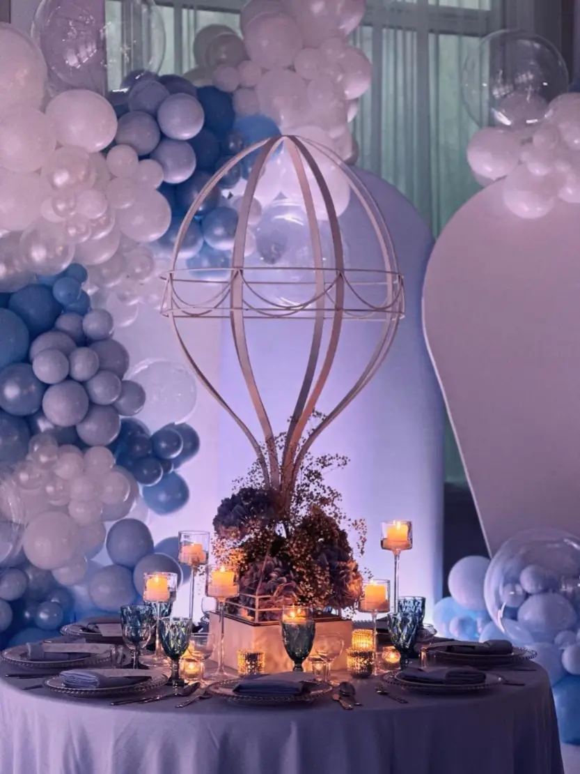 Elegancka dekoracja stołu z motywem balonowym, idealna na przyjęcie dla dzieci lub baby shower. Centralnym punktem jest dekoracyjna rzeźba w kształcie balonu na gorące powietrze, otoczona kompozycją z fioletowych hortensji, białej gipsówki i podświetlona świecami w szklanych świecznikach. Tło stanowi imponująca aranżacja z białych i niebieskich balonów w różnych odcieniach i rozmiarach. Na stole znajdują się talerze z dekoracyjnymi obrzeżami, niebieskie kieliszki oraz srebrne sztućce, tworząc magiczną i baśniową atmosferę