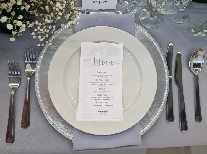 Na elegancko nakrytym stole znajduje się talerz z ozdobnym, srebrnym brzegiem, na którym leży menu zawierające przystawkę, danie główne i deser. Menu jest starannie złożone na szarym serwetniku, który jest ozdobiony delikatnym, szarym materiałem. W tle widać przezroczyste kieliszki i szklanki, a także subtelną kompozycję z białymi kwiatkami, które dekorują stół. Wokół talerza rozmieszczono sztućce, w tym widelce, noże i łyżki, tworząc estetyczną i elegancką aranżację