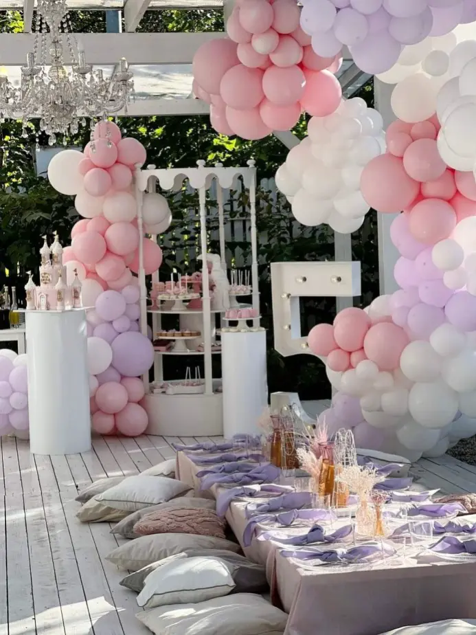 Plenerowa aranżacja przyjęcia w stylu boho glamour z dominacją pastelowych odcieni różu, fioletu i bieli. W centralnym punkcie znajduje się biały, okrągły candy bar z dekoracją balonową w różnych rozmiarach oraz girlandy balonowe otaczające całą strefę słodkości. Na podłodze z bielonego drewna rozłożono poduszki i ustawiono niski stół z lawendowymi serwetkami, szklanymi nakryciami i butelkami z suszonymi trawami pampasowymi. Dekorację dopełnia efektowny żyrandol oraz duża litera z podświetleniem LED w tle