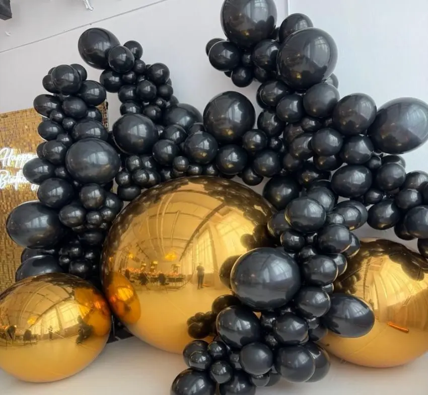 Dekoracja z balonów na urodziny lub inną okazję, charakteryzująca się elegancką kombinacją czarnych i złotych balonów. Balony w różnych rozmiarach tworzą dynamiczną, nowoczesną kompozycję, która wyróżnia się dużymi, połyskującymi złotymi kulami w centrum, otoczonymi mniejszymi czarnymi balonami. Tego rodzaju aranżacja balonowa może być doskonałą ozdobą stołu, fotobudki lub jako tło do sesji zdjęciowej. W tle widoczna jest również dekoracja z napisem Happy Birthday, co sugeruje, że jest to część przestrzeni z okazji urodzin