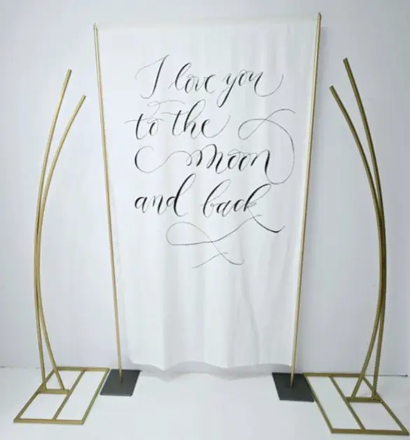 Stylowa dekoracja z napisem I love you to the moon and back, która z pewnością doda uroku romantycznemu wydarzeniu. Biała tkanina zawieszona na eleganckich, złotych ramach, przyciąga wzrok pięknym, kaligraficznym pismem. Napis, wyrażający miłość i oddanie, świetnie sprawdzi się podczas wesel, rocznic, sesji zdjęciowych czy innych wyjątkowych okazji, które celebrują miłość. Złote ramy stanowią nowoczesny akcent, doskonale łącząc romantyzm z eleganckim designem