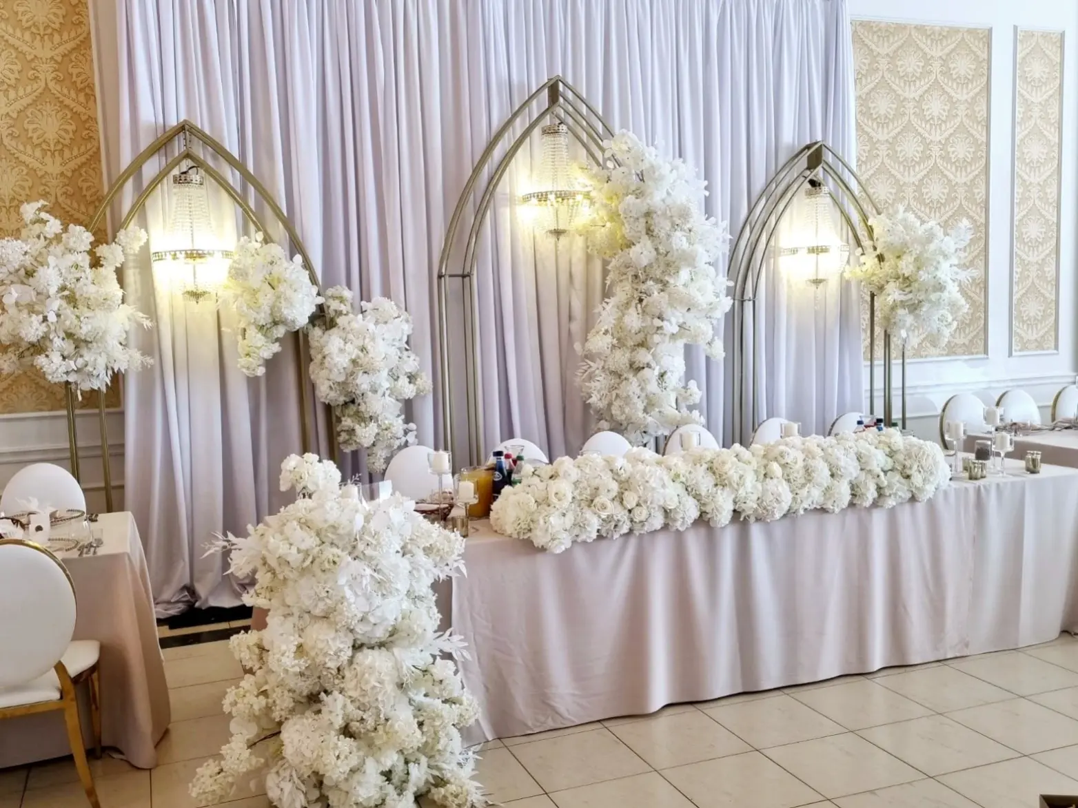 Elegancka aranżacja sali weselnej lub eventowej z ozdobami w odcieniach bieli i złota. Trzy wysokie łukowe konstrukcje, ozdobione obficie białymi kwiatami, tworzą przepiękny dekoracyjny punkt na tle delikatnej białej tkaniny. Na stoliku głównym umieszczono bukiety kwiatów, a całość otacza harmonijnie uporządkowane kompozycje kwiatowe na stole. Przestrzeń jest rozświetlona przez stylowe żyrandole w kształcie kul, które doskonale komponują się z architekturą sali. Białe krzesła i eleganckie obrusy dopełniają wrażenie luksusu i klasycznego piękna