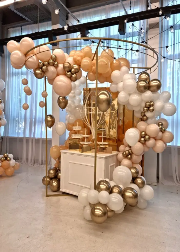 Elegancka dekoracja balonowa w odcieniach bieli, złota, brzoskwini i pudrowego różu, tworząca efektowną strefę na przyjęcie. Konstrukcja z cienkich, złotych obręczy otacza biały stolik z ciastkami i słodkościami, ustawiony na tle złotej, lustrzanej ścianki. Balony w różnych rozmiarach tworzą gęste girlandy, oplatające całość. Na górze i po bokach zawieszone są także złote balony w metalicznym połysku. Całość znajduje się w jasnym, przestronnym wnętrzu z delikatnymi zasłonami i girlandami świetlnymi. Dekoracja idealna na urodziny, baby shower lub eleganckie przyjęcie