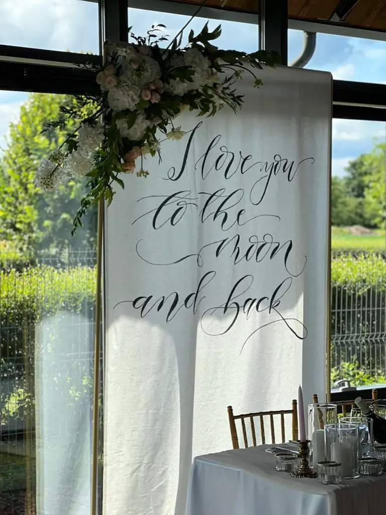 Romantyczna dekoracja ślubna z białym banerem z kaligraficznym napisem „I love you to the moon and back”, zawieszonym w oknie z widokiem na ogród. Górna część materiału ozdobiona jest kompozycją z białych i pudroworóżowych kwiatów, zieleni oraz drobnych akcentów suszonych roślin. Obok ustawiony jest stół z białym obrusem, świecami w szklanych świecznikach i elegancką zastawą. Całość tworzy intymną i nastrojową przestrzeń idealną na przyjęcie weselne, sesję zdjęciową lub strefę nowożeńców