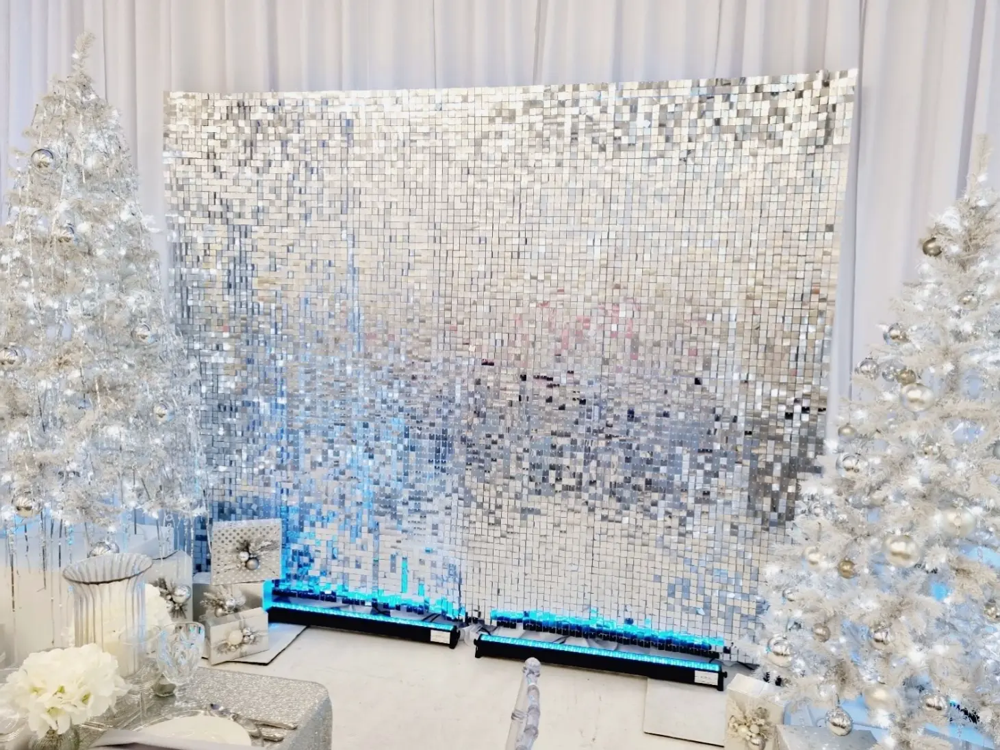 Zimowa dekoracja eventowa w stylu glamour, z centralnie umieszczoną srebrną ścianką shimmer wall z lustrzanych paneli odbijających światło. Po obu stronach ustawiono białe choinki udekorowane srebrnymi bombkami i światełkami, tworząc elegancki, świąteczny klimat. Na pierwszym planie widoczne są ozdobnie zapakowane prezenty, kryształowe naczynia i biała kwiatowa kompozycja. Całość podświetlona subtelnym niebieskim światłem od dołu, idealna jako tło do sesji zdjęciowych, świątecznych eventów firmowych lub luksusowego przyjęcia bożonarodzeniowego.