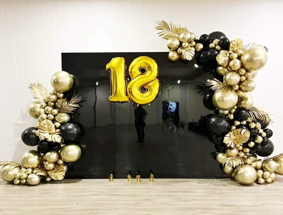 Dekoracja urodzinowa na 18. urodziny w eleganckim stylu glamour. Centralnym elementem jest czarna błyszcząca ścianka z balonowymi cyframi „18” w kolorze metalicznego złota. Po bokach znajdują się efektowne girlandy z balonów w odcieniach czerni i złota, uzupełnione złotymi liśćmi tropikalnymi dla luksusowego efektu. Całość osadzona na jasnym panelu podłogowym, tworzy idealne tło do zdjęć i celebracji pełnoletniości