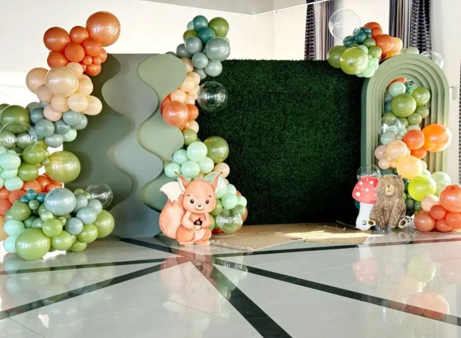 Dekoracja w leśnym stylu z balonami w pastelowych odcieniach zieleni, pomarańczu, błękitu i beżu, idealna na przyjęcie dla dzieci lub baby shower. Tło stanowi zielona ściana z imitacją żywopłotu oraz geometryczne panele w kształcie fali i łuku. U podstawy dekoracji znajdują się urocze postacie zwierząt: wiewiórka i miś, a także grzybki. Przezroczyste balony dodają lekkości, a całość tworzy bajkowy klimat natury i lasu