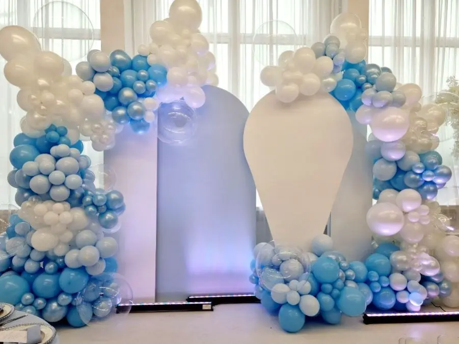 Dekoracja eventowa w pastelowych odcieniach błękitu i bieli, idealna na baby shower lub urodziny chłopca. Tło składa się z trzech paneli w kształcie balonów i łuków, w kolorze białym i jasnoniebieskim. Całość ozdobiona jest efektownymi girlandami balonowymi w odcieniach błękitu, bieli i perłowego srebra. W tle znajdują się białe firany przepuszczające światło, co nadaje aranżacji elegancki i delikatny charakter