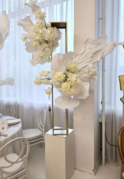 owoczesna dekoracja florystyczna w stylu minimalistycznym, przedstawiająca elegancką kompozycję z białych sztucznych kwiatów — róż, hortensji i dużych, dekoracyjnych liści — umieszczonych w geometrycznym stojaku na wysokim, białym cokole. Całość utrzymana w bieli i złocie, ustawiona przy oknie z białymi firanami, obok białych krzeseł i stołów. Idealna dekoracja na wesele, bankiet, chrzest, komunię lub nowoczesne przyjęcie glamour