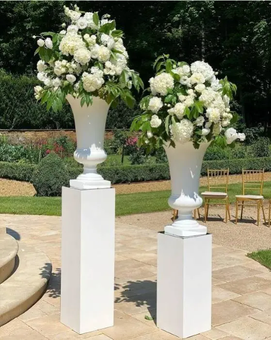 Eleganckie dekoracje kwiatowe w wysokich białych wazonach ustawione na białych postumentach w plenerze, idealne na ceremonię ślubną w ogrodzie. Kompozycje składają się z bujnych, białych hortensji, róż i piwonii oraz dużych, zielonych liści, tworząc świeży, klasyczny i romantyczny efekt. W tle widać ogrodowe alejki, zieleń oraz złote krzesła ustawione dla gości