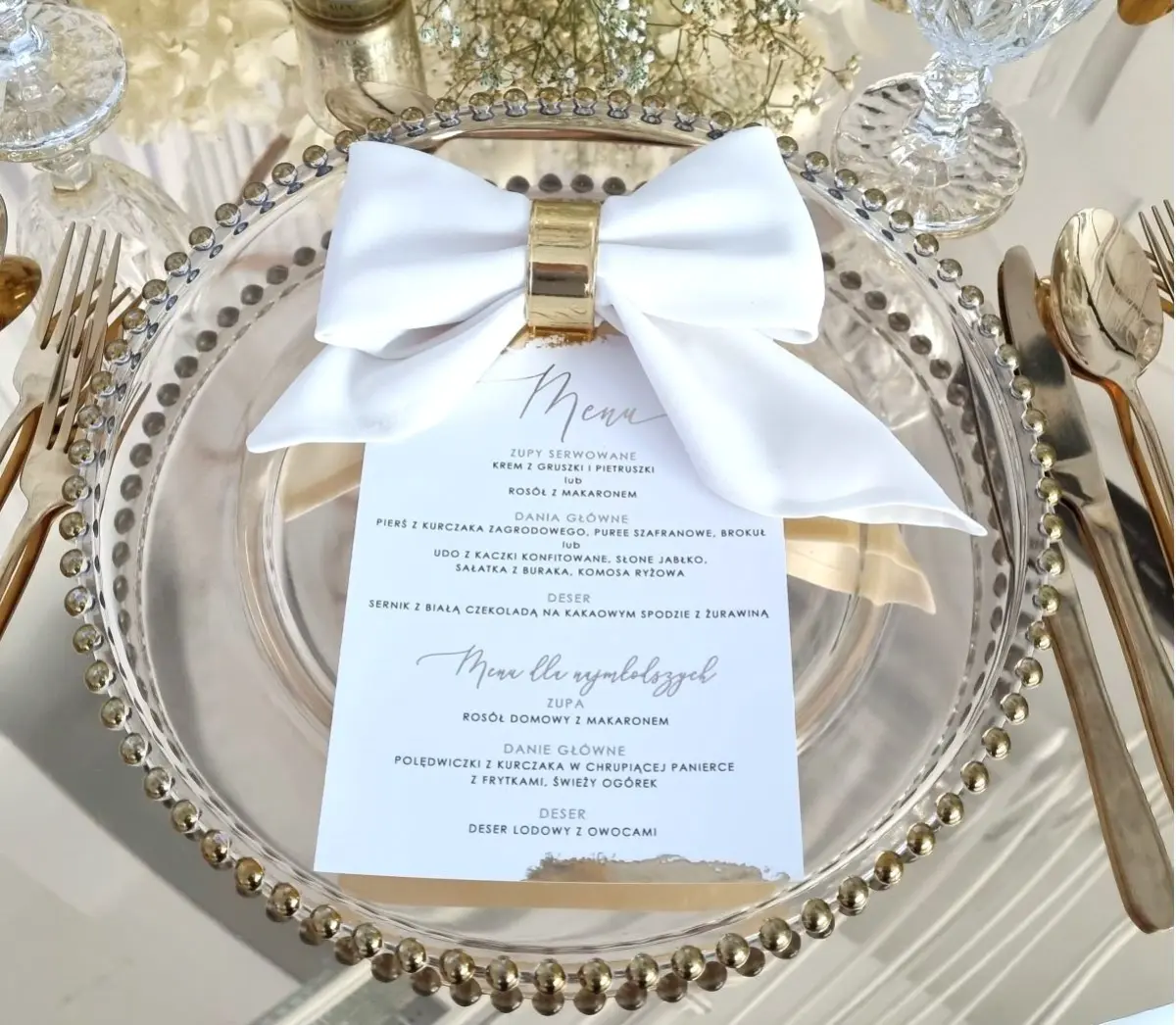 Na zdjęciu znajduje się eleganckie nakrycie stołowe. W centrum widoczny jest przezroczysty talerz ozdobiony złotą obręczą. Na talerzu znajduje się karta menu z elegancką, odręcznie wyglądającą czcionką, a obok niej leży biała serwetka, złożona w kształt kokardy i spięta złotym pierścieniem. Po prawej stronie talerza widać złotą łyżkę i nóż, a po lewej stronie dwa widelce. W tle widoczne są kieliszki do wina, a całość otoczona jest subtelnymi, złotymi akcentami, które dodają całości elegancji. Tło to białe kwiaty, co podkreśla wyrafinowany, romantyczny charakter nakrycia