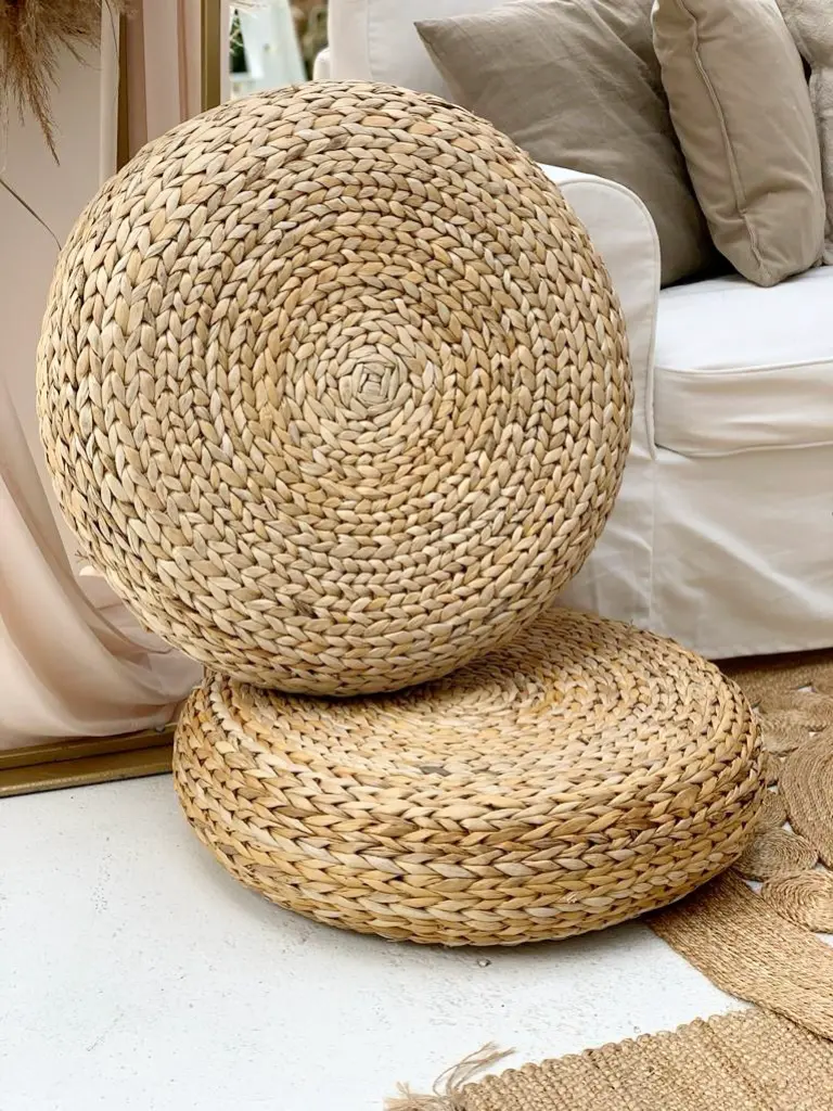 Zbliżenie na dwa okrągłe pufy plecione z naturalnej trawy morskiej w stylu boho, ustawione jeden na drugim. W tle widoczna biała sofa z beżowymi poduszkami oraz fragmenty jutowych dywanów i jasnej zasłony. Naturalna kolorystyka i tekstura puf wpisują się w ekologiczny, rustykalny wystrój wnętrza lub eventu. Idealny dodatek do aranżacji ślubnych, przyjęć plenerowych lub stref chillout w stylu boho i eko