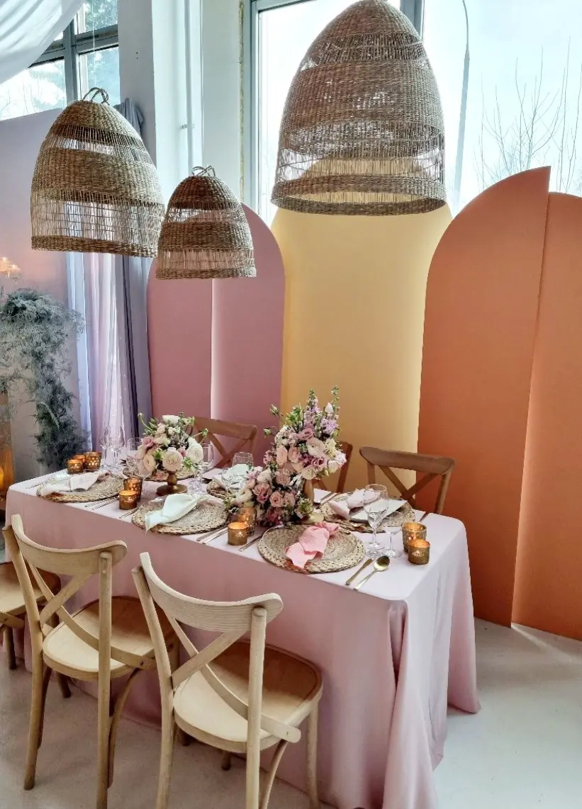 Kolorowa aranżacja stołu w stylu boho z pastelowymi akcentami i naturalnymi materiałami. Stół nakryty różowym obrusem, ozdobiony plecionymi podkładkami, złotymi sztućcami, białymi i różowymi serwetkami oraz kompozycjami kwiatowymi z róż i eustomy w odcieniach kremu, różu i fioletu. Ciepłe światło świec w złotych szklanych lampionach tworzy przytulny klimat. W tle dekoracyjne panele w kształcie łuków w pastelowych kolorach – brzoskwiniowym, morelowym i różowym. Nad stołem zawieszone wiklinowe lampy, podkreślające naturalny, rustykalny styl idealny na wesele lub garden party