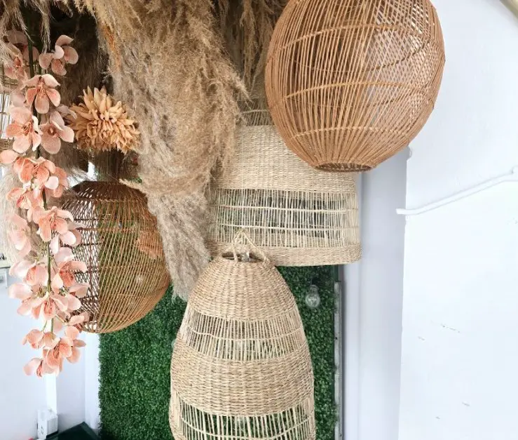 Dekoracja sufitowa w stylu boho złożona z plecionych, naturalnych kloszy lamp w różnych kształtach i odcieniach beżu i brązu. Wśród nich znajdują się kuliste i stożkowe formy, zawieszone w grupie. Towarzyszą im suszone trawy pampasowe i kwiaty w odcieniach brzoskwini i różu, tworzące efektowną kompozycję. W tle zielona ściana z roślin oraz białe elementy architektoniczne. Idealna dekoracja na ślub, garden party lub sesję zdjęciową w stylu boho, rustykalnym i eko