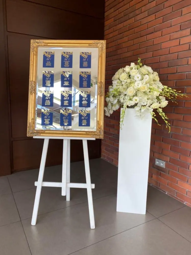 Elegancka tablica stołów weselnych w złotej ramie na białej sztaludze, z granatowymi kartami oznaczonymi numerami stołów i listą gości, ozdobionymi złotymi kwiatowymi ornamentami. Obok dekoracyjna kompozycja kwiatowa z białych róż, eustom i hortensji w wysokim, białym postumencie. Ustawione przy ścianie z czerwonej cegły, tworzą stylową strefę powitalną