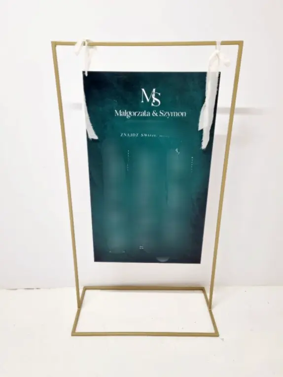 Minimalistyczna tablica z rozkładem stołów weselnych w eleganckiej złotej ramie stojącej na podłodze. Ciemnozielona karta z imionami Małgorzata & Szymon i napisem Znajdź swoje miejsce zawieszona jest na białych tasiemkach. Nowoczesny design i czytelny układ wpisują się w styl glamour oraz industrial chic. Idealna dekoracja na eleganckie wesele